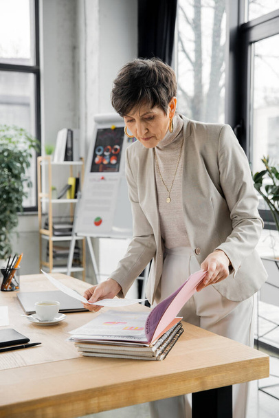femme d'affaires moderne d'âge moyen regardant des documents près de tasse de café dans le bureau, paperasse - Photo, image