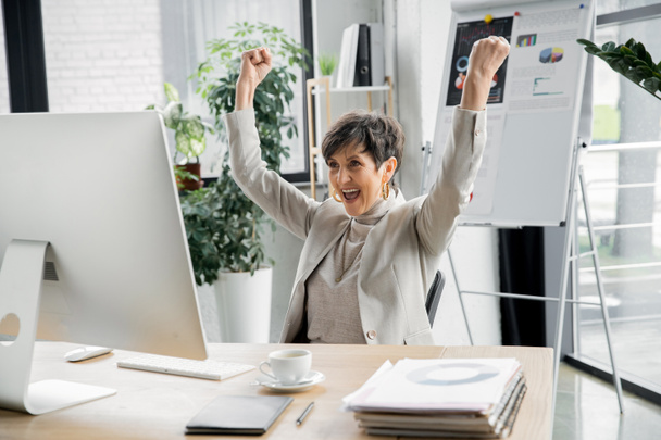 excited entrepreneur showing win gesture near computer monitor in office, business success - Foto, Imagen