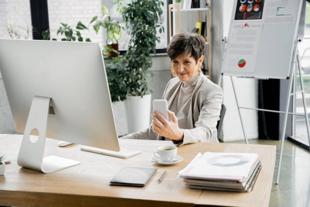 счастливая зрелая деловая женщина смотрит на смартфон рядом с компьютером, ноутбуки и документы на рабочем месте - Фото, изображение