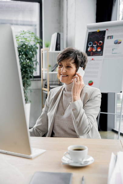 радостная деловая женщина средних лет разговаривает на смартфоне возле чашки кофе в современном офисе - Фото, изображение
