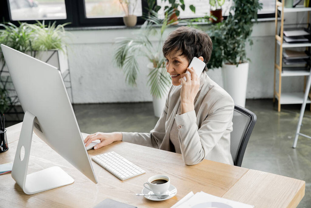 Lächelnder Manager mittleren Alters im Gespräch mit dem Handy in der Nähe von Laptop und Kaffee im modernen Büro - Foto, Bild