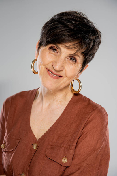 brunette femme d'âge moyen en veste brune et boucles d'oreilles cerceau souriant à la caméra sur gris, mode automne - Photo, image