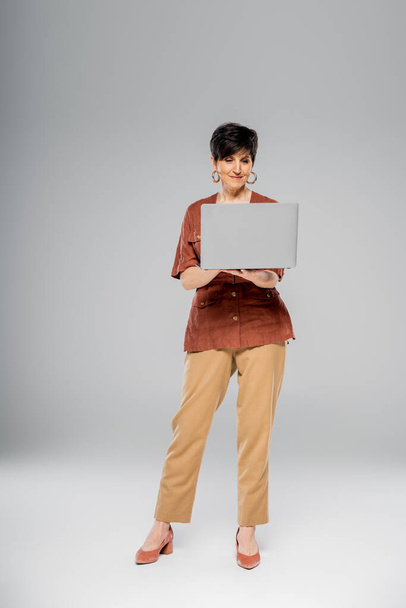 Dojrzałe businesswoman w brązowej kurtce przy użyciu laptopa na szary, jesień, sezonowy strój, trend mody - Zdjęcie, obraz