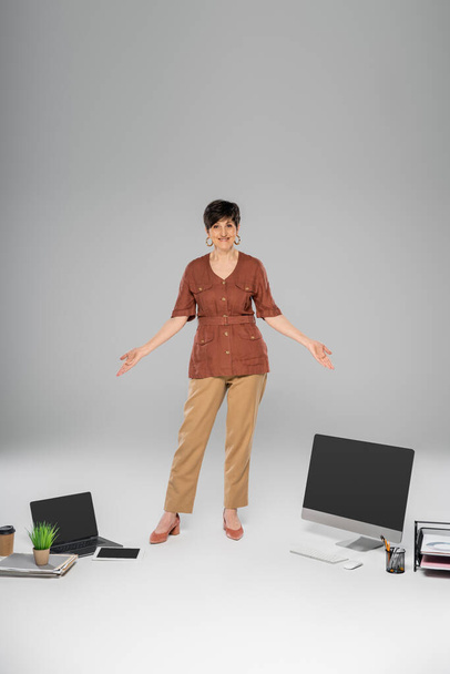 зрелая предпринимательница в осеннем наряде, указывающая на цифровые устройства, документы и бумажную чашку на сером - Фото, изображение