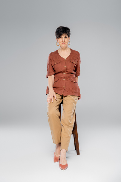 Осенняя мода, женщина средних лет в модном наряде сидит на высоком стуле на сером, полной длины - Фото, изображение