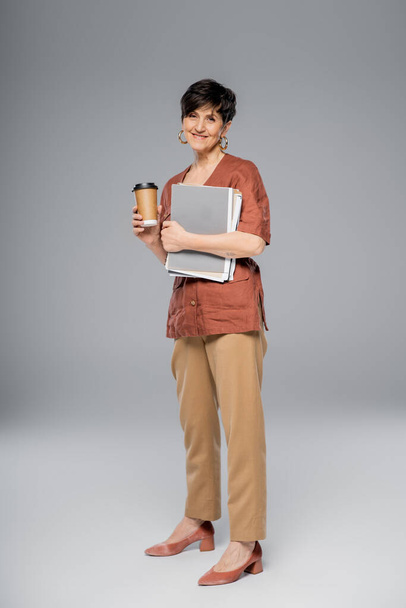 trendy middle aged businesswoman with coffee to go and documents on grey, autumn fashion trend - Photo, Image