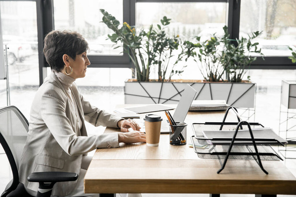 Seitenansicht einer selbstbewussten Geschäftsfrau mittleren Alters, die in der Nähe eines Imbisses im modernen Büro am Laptop arbeitet - Foto, Bild
