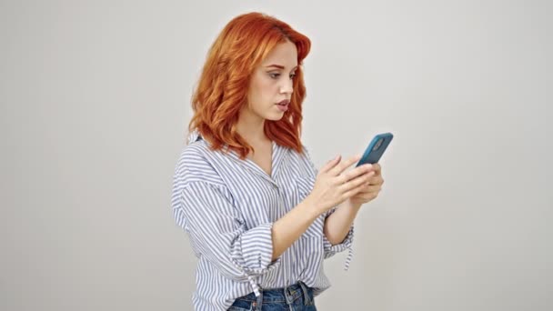 Nuori punapää nainen seisoo yllätys ilme käyttäen älypuhelin yli eristetty valkoinen tausta - Materiaali, video