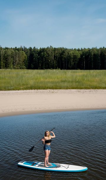 Молодая стройная женщина в летнее время гребля на доске SUP на красивом озере, здоровый образ жизни - Фото, изображение