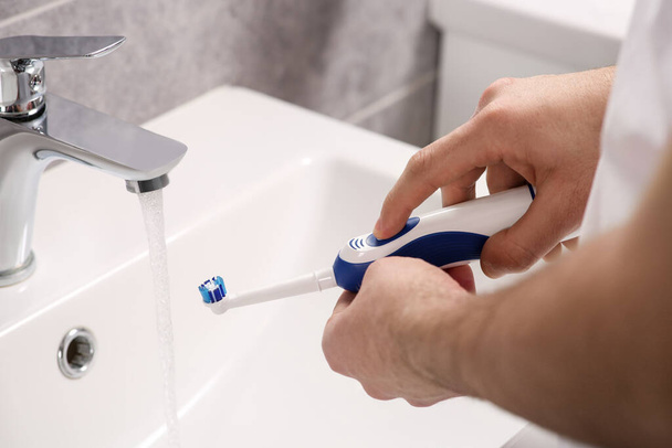 Man holding electric toothbrush near flowing water above sink in bathroom, closeup - Foto, Imagen
