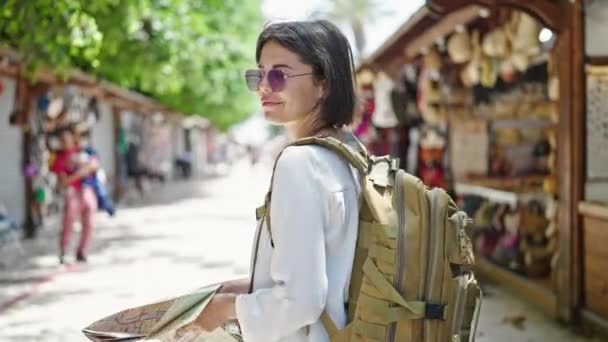 Joven hermosa mujer hispana turista sonriendo confiado sosteniendo mapa de la ciudad en el mercado callejero - Metraje, vídeo