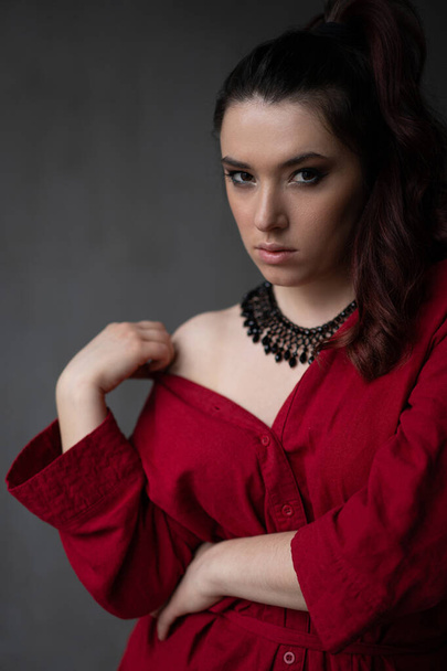 a beautiful young girl in a red shirt with a necklace with a handmade ornament on her neck is posing in a photo studio - Photo, Image