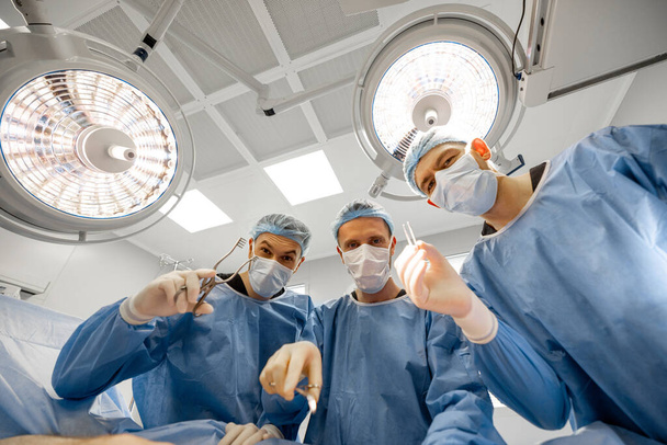 Portrét tří děsivých chirurgů s chirurgickým nářadím se dívá do kamery na operačním sále - Fotografie, Obrázek