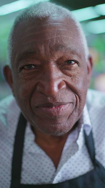 Close-up rosto de um idoso afro-americano sênior com cabelos grisalhos e rugas vestindo um avental preto olhando para câmera com expressão neutra - Foto, Imagem