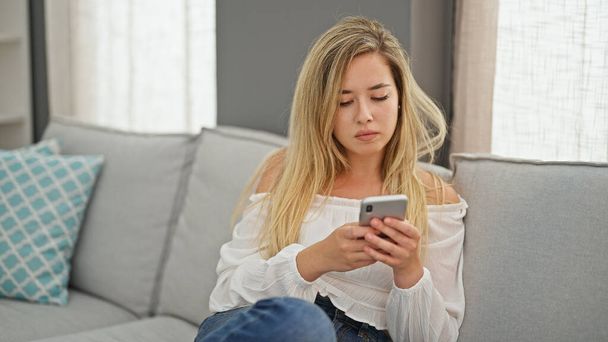 Young blonde woman using smartphone sitting on sofa thinking at home - Photo, Image