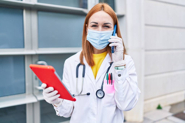 Tıbbi maske takan, dokunmatik ped kullanan beyaz kadın doktor hastanede akıllı telefondan konuşuyor. - Fotoğraf, Görsel