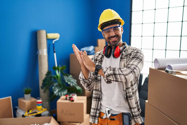Молодой латиноамериканец с бородой, работающий над ремонтом дома хлопает и аплодирует счастливым и радостным, улыбающимся гордым рукам вместе  - Фото, изображение