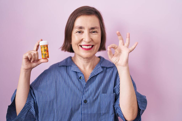 Mujer hispana de mediana edad sosteniendo píldoras haciendo signo bien con los dedos, sonriendo gesto amistoso excelente símbolo  - Foto, imagen