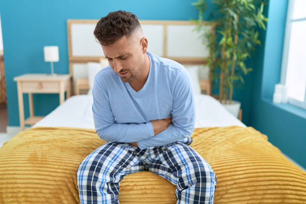 Молодий іспаномовний чоловік страждає від болю в животі, сидячи в ліжку в спальні
 - Фото, зображення