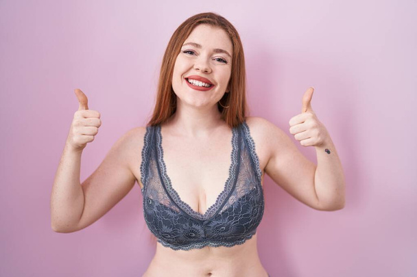 Redhead woman wearing lingerie over pink background success sign doing positive gesture with hand, thumbs up smiling and happy. cheerful expression and winner gesture.  - Photo, Image