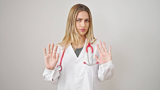 Joven mujer rubia médico de pie con expresión seria haciendo stop gesture sobre fondo blanco aislado - Foto, imagen