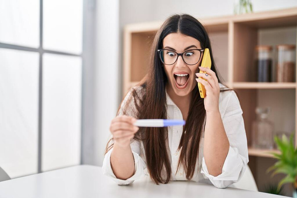 Mujer morena joven sosteniendo el resultado de la prueba de embarazo hablando por teléfono celebrando loco y sorprendido por el éxito con los ojos abiertos gritando emocionado.  - Foto, imagen