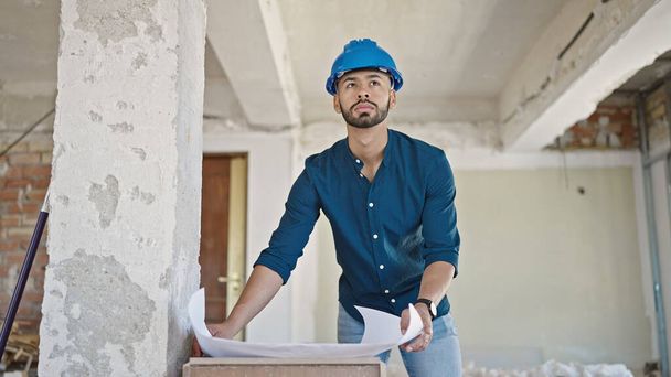 Young hispanic man architect wearing hardhat looking at blueprints at construction site - Photo, Image