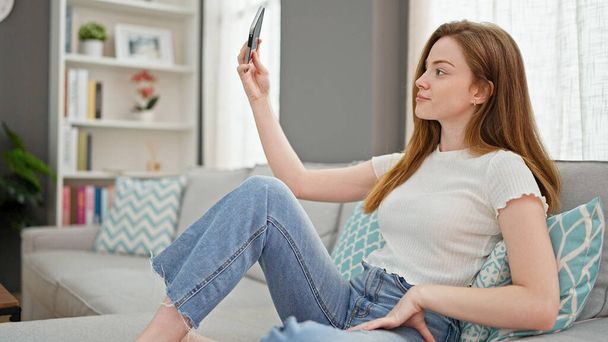 Молода блондинка знімає селфі з смартфоном, що сидить на дивані вдома
 - Фото, зображення