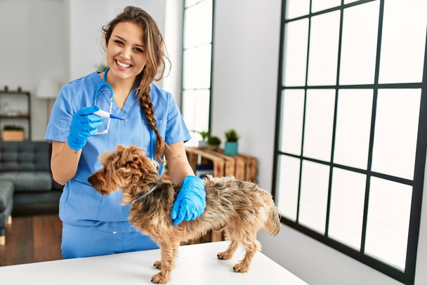 Young beautiful hispanic woman veterinarian measuring dog temperature holding thermometer at home - Photo, Image