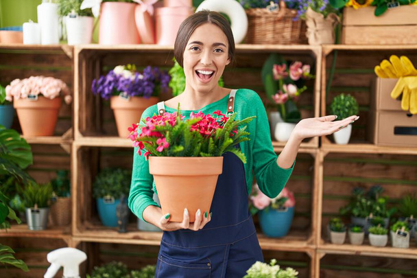 Młoda Latynoska kobieta pracuje w kwiaciarni gospodarstwa roślin świętując osiągnięcie z uśmiechem i szczęśliwy wyraz zwycięzcy z podniesioną ręką  - Zdjęcie, obraz