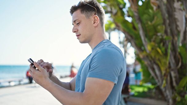 Joven turista hispano usando smartphone con expresión relajada a orillas del mar - Foto, imagen