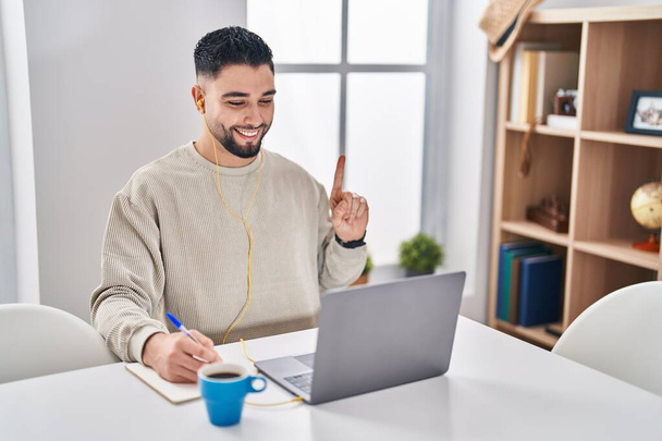Νεαρός όμορφος άντρας που χρησιμοποιεί φορητό υπολογιστή κάνει online κλήση χαμογελώντας με μια ιδέα ή ερώτηση δείχνοντας το δάχτυλο με χαρούμενο πρόσωπο, νούμερο ένα  - Φωτογραφία, εικόνα