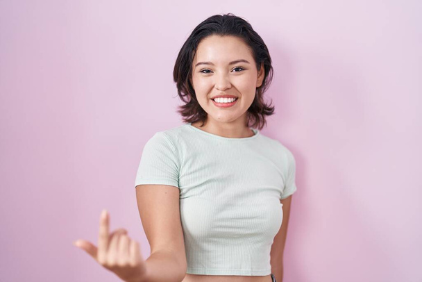 Ισπανίδα νεαρή γυναίκα στέκεται πάνω από ροζ φόντο beckoning έρθει εδώ χειρονομία με το χέρι καλώντας φιλόξενο ευτυχισμένη και χαμογελώντας  - Φωτογραφία, εικόνα