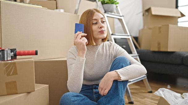 Jovem loira sentada no chão pensando segurando cartão de crédito na nova casa - Foto, Imagem