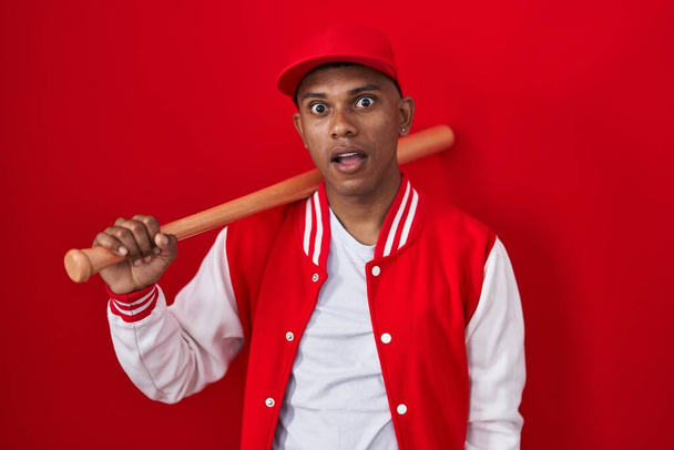 Young hispanic man playing baseball holding bat in shock face, looking skeptical and sarcastic, surprised with open mouth  - Photo, Image