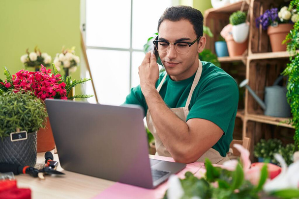 Молодий іспаномовний чоловік флорист розмовляє на смартфоні за допомогою ноутбука в квітковому магазині
 - Фото, зображення