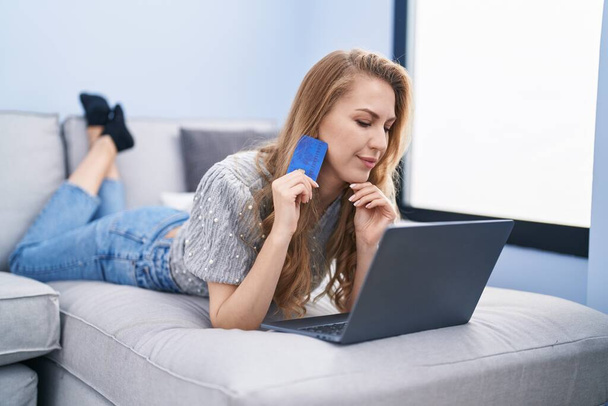 Mladá blondýna žena pomocí notebooku a kreditní karty ležící na pohovce doma - Fotografie, Obrázek