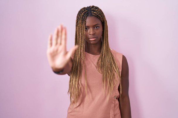 Αφροαμερικανή γυναίκα με πλεγμένα μαλλιά που στέκεται πάνω από ροζ φόντο κάνει σταματήσει να τραγουδά με την παλάμη του χεριού. προειδοποιητική έκφραση με αρνητική και σοβαρή χειρονομία στο πρόσωπο.  - Φωτογραφία, εικόνα