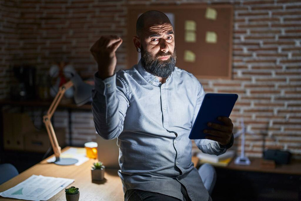 Młody Hiszpan z brodą i tatuażami, pracujący w biurze w nocy, wykonujący włoski gest z ręką i palcami.  - Zdjęcie, obraz