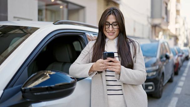 Giovane bella donna ispanica utilizzando smartphone appoggiato sulla macchina in strada - Foto, immagini