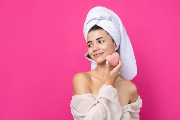 Όμορφη χαρούμενη ελκυστική κοπέλα με μια πετσέτα στο κεφάλι της, κρατά ένα σφουγγάρι σε μορφή ροζ καρδιάς. - Φωτογραφία, εικόνα