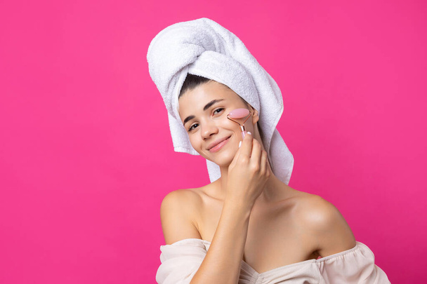 Una bella ragazza attraente allegra con un asciugamano sulla testa tiene un rullo cosmetico vicino al suo viso su uno sfondo rosa. - Foto, immagini