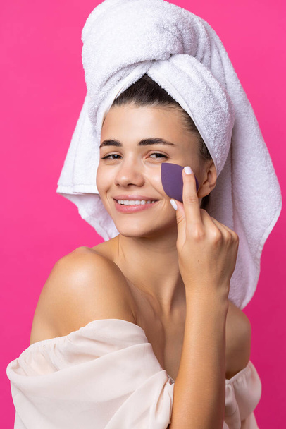 Beautiful cheerful attractive girl with a towel on her head, holds a sponge against a pink background. - Foto, Imagem