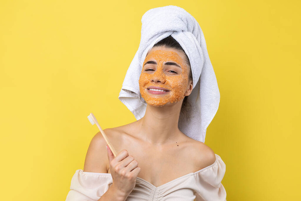 A cute young girl with a scrub mask on her face brushes her teeth against a yellow background. - Valokuva, kuva