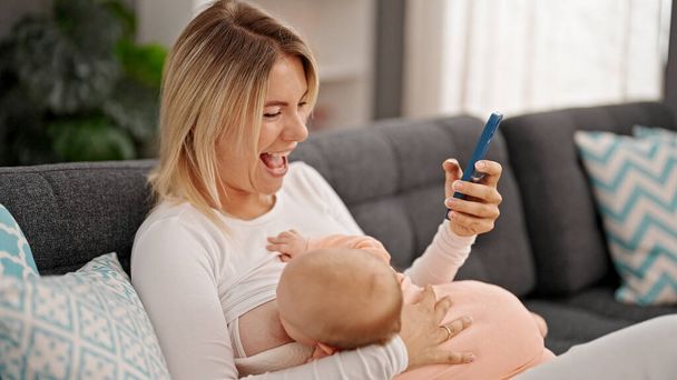 Mutter und Tochter sitzen zu Hause mit Smartphone auf Sofa und stillen Baby - Foto, Bild