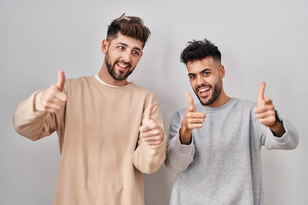 Jeune couple homosexuel debout sur fond blanc pointant les doigts vers la caméra avec un visage heureux et drôle. bonne énergie et vibes.  - Photo, image