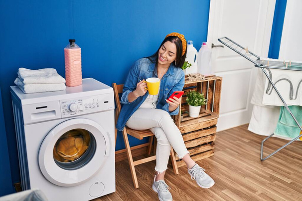 Junge schöne hispanische Frau trinkt mit Smartphone Kaffee und wartet auf Waschmaschine in der Waschküche - Foto, Bild