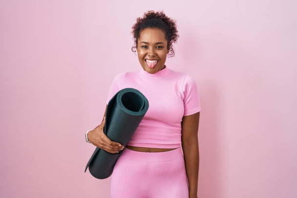 Mulher hispânica jovem com cabelo encaracolado segurando tapete de ioga sobre fundo rosa colando a língua para fora feliz com expressão engraçada. conceito de emoção.  - Foto, Imagem