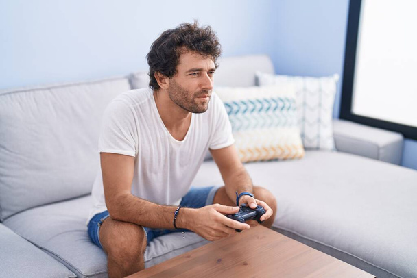 Young hispanic man playing video game sitting on sofa at home - Фото, изображение