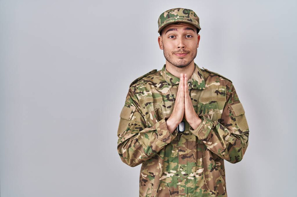 Jeune homme hispanique portant l'uniforme de l'armée de camouflage priant avec les mains ensemble demandant pardon souriant confiant.  - Photo, image